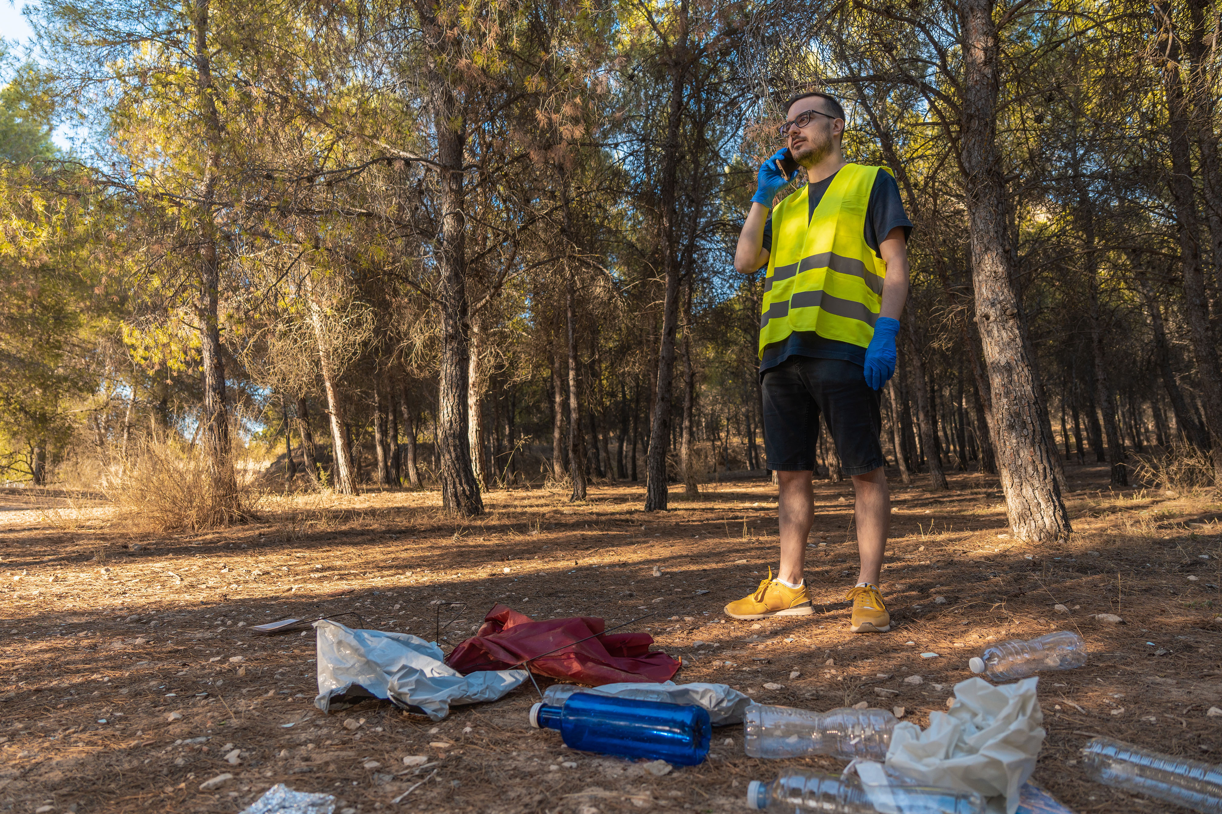In einem Waldstück steht ein Mann in gelber Warnweste über einen Haufen vom verstreutem Müll und redet am Smartphone.