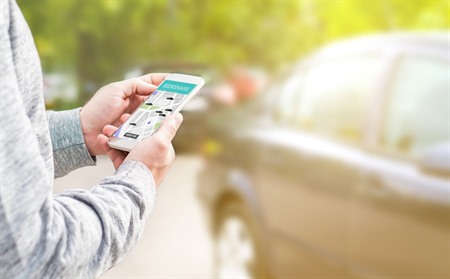 Carsharing: Eine Person hält ein Smartphone in der Hand; im Hintergrund steht ein Auto