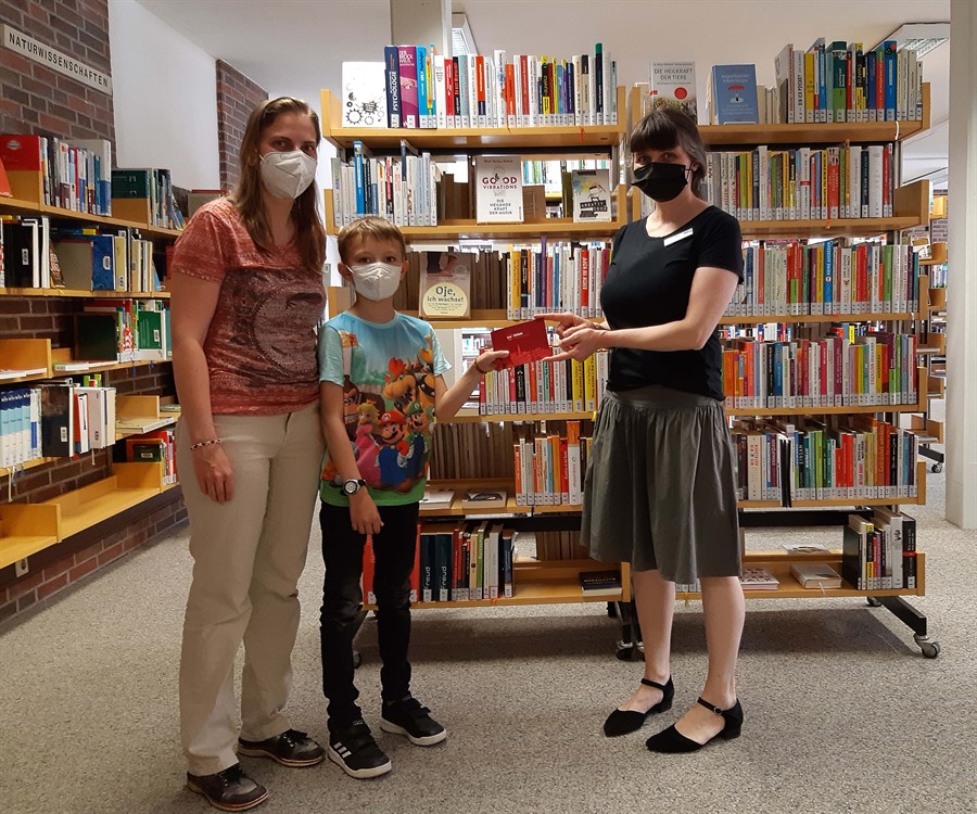 Vor einem Bücherregal übergibt Christine Daum, Mitarbeiterin der Stadtbücherei, einen Stadtgutschein an Kerstin Hasse und ihren Sohn Henry.