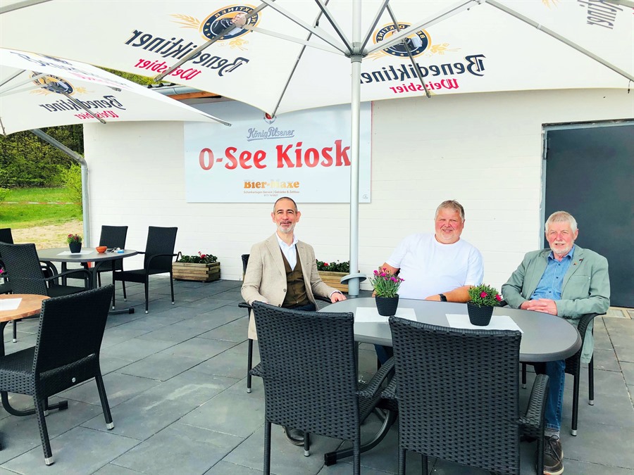 Stadtbaurat Dr. Andreas Stefansky, Pächter Matthias Neumann und Klaus Knust, Ortsbürgermeister von Oldenstadt, sitzen an einem Tisch auf der frisch gepflasterten Terrasse unter einem Sonnenschirm.