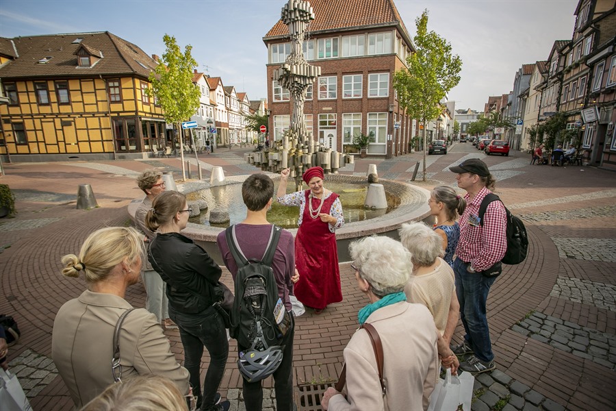 Eine Gästeführerin im mittelalterlichem Gewand steht vor dem Brunnen auf dem Schnellenmarkt und erzählt einer Gruppe von Menschen Interessantes über die Hansezeit.