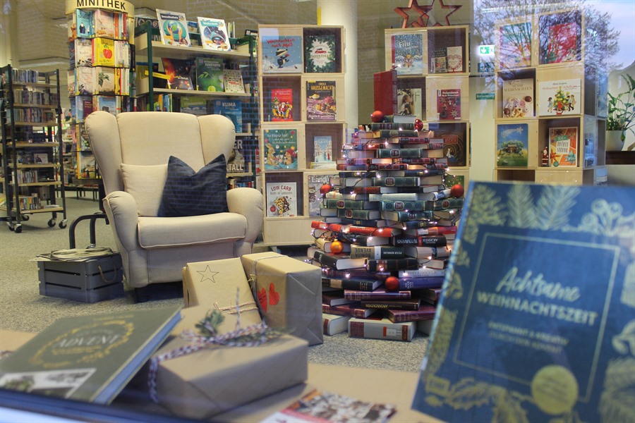 Ein weißer Sessel steht vor Bücherregalen. Vor dem Sessel sind Bücher in Form eines Weihnachtsbaums gestapelt und mit einer Lichterkette geschmückt.  