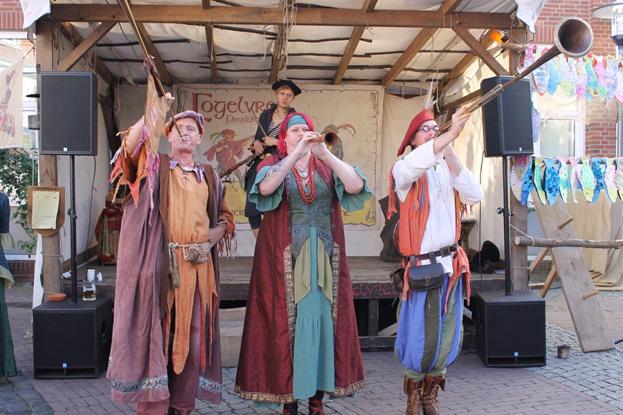Drei Musikanten in mitteralterlichen Gewänden stehen vor einer Bühne und bedienen verschiedene Blasinstrumente.