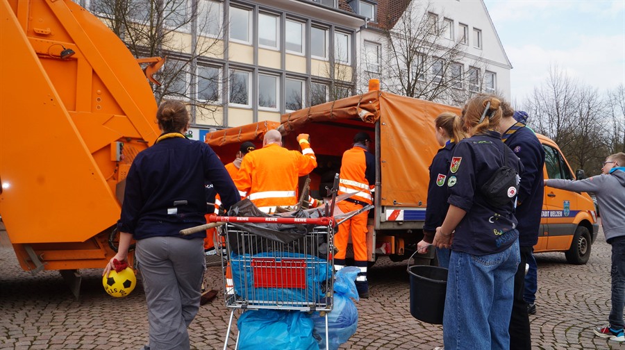 Ein Einkaufswagen voll mit Unrat wird von drei Personen zu zwei stehenden, orangefarbenen Abfallentsorgungsfahrzeugen geschoben.