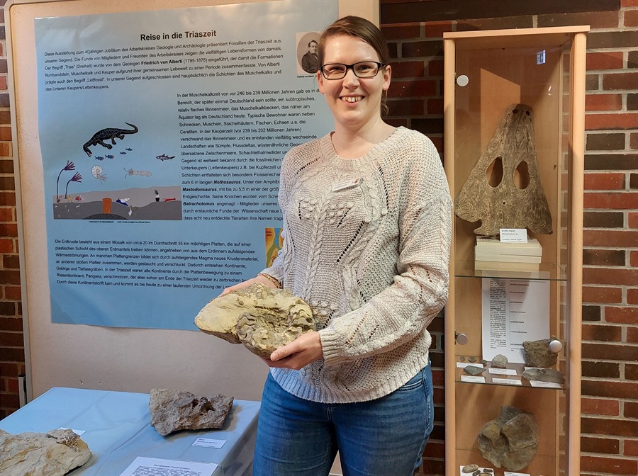 Eine Frau hält eine Fossilie in der Hand und steht vor einem Plakat mit Informationen über Saurier und Schaukästen mit weiteren Fossilien..