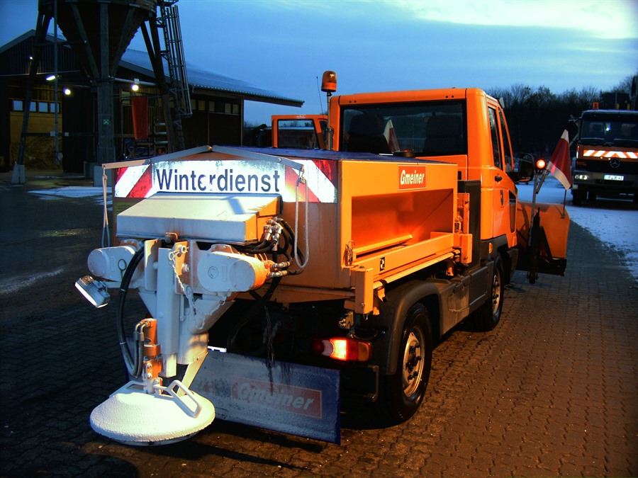 Ein orangenes Winterdienst-Fahrzeug in Aktion: Auf der Heckklappe steht "Winterdienst".